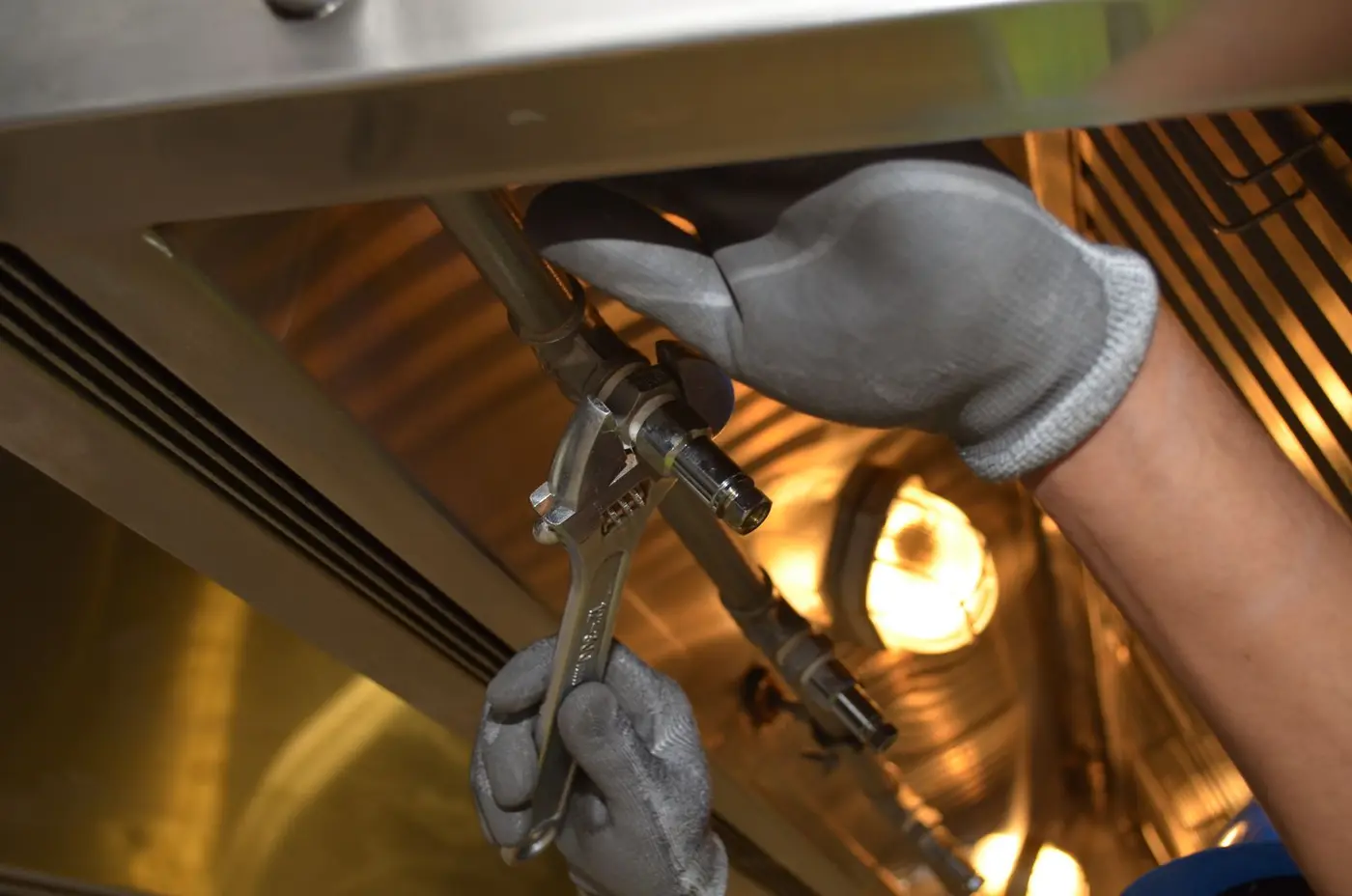 Gloved hands tightening a fitting on an overhead pipe with an adjustable wrench