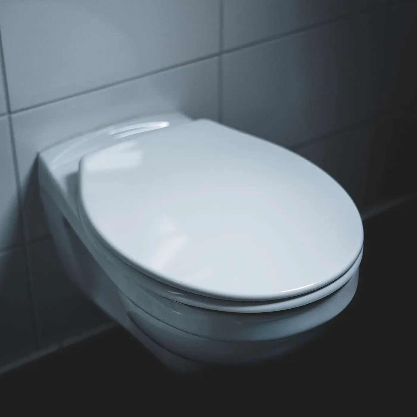 A white wall mounted toilet against gray tile in a dim bathroom