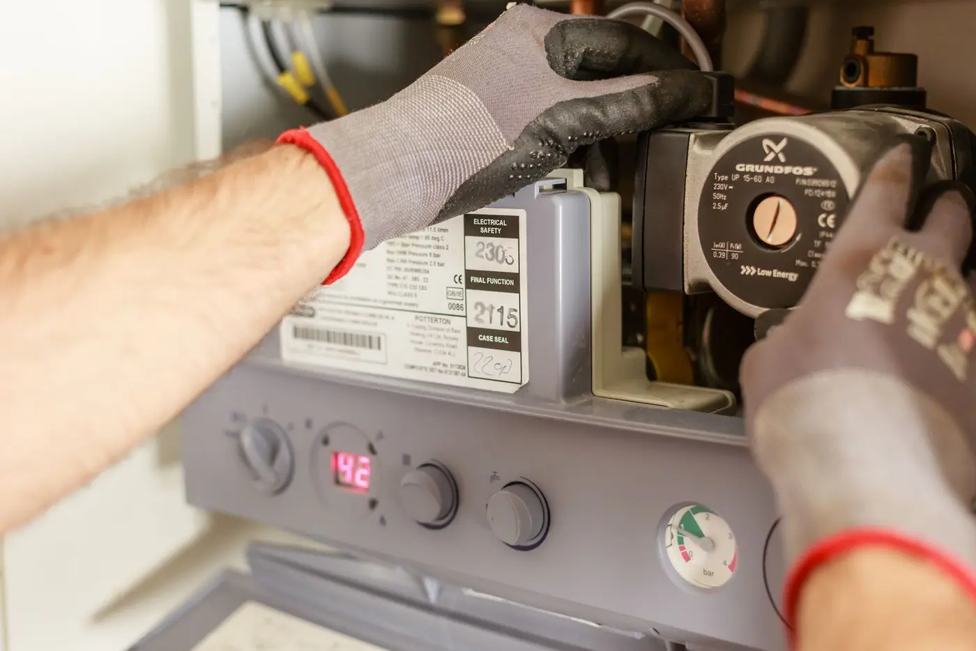 Gloved hands servicing the open internals of a water heater, with the circulator pump and pressure gauge visible